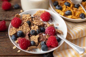 Cornflakes with blueberries and raspberries in white bowl on dark wooden desk.