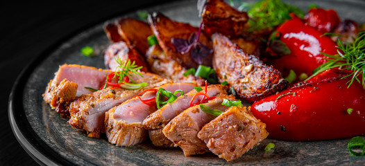 fried meat with potatoes, pepper, tomatoes, herbs and spices in plate on black wooden table background