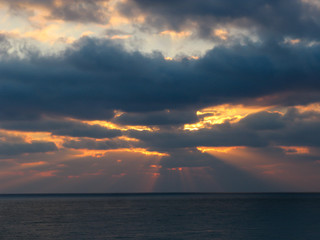 Naklejka na ściany i meble bright rays of the sun at dawn at sea through dark clouds