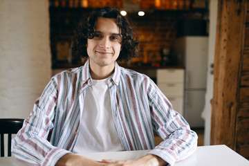 People and lifestyle concept. Portrait of handsome cheerful young man with curly black hair and bristle sitting at white table indoors, staring at camera with happy smile, having friendly look