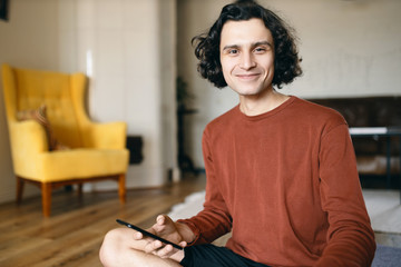 Happy young male with black wavy hair enjoying online communication using high speed wireless internet connection on smart phone, texting girlfriend, sitting in stylish living room interior