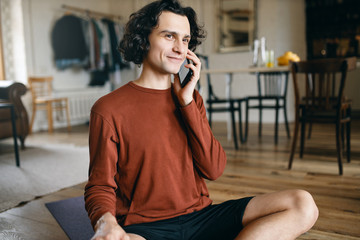 Happy young male in casual clothes spending all day at home alone while social distancing, smiling, glad to hear voice of his girlfriend, having phone conversation, sharing news. Electronic gadgets