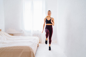Strong Athletic Fit girl in top and leggings is Energetically Jogging and jumping with smile in Place at Home in Her Spacious and Bright bedroom with Minimalistic white interior.