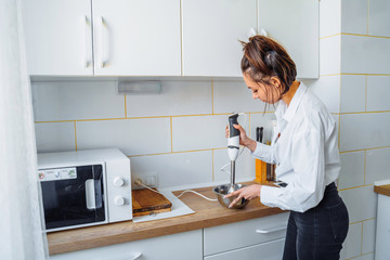 Attractive girl preparing dough in a food processor while standing in a bright kitchen. Mixing dough with hand mixer on kitchen table background with cooking ingredients