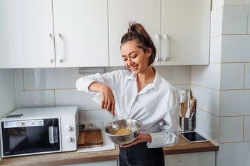 Beautiful, attractive, smiling young woman kneading dough with whisk for banana pancakes in steel...