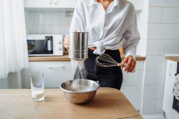 DIY home cooking concept. A bright brunette woman sifting the flour for pancakes through a sieve, salting the dough, adding milk. Young woman smiling, loves to cook in a beautiful modern kitchen.