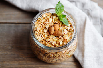 Homemade oatmeal granola in an open glass jar, decorated with nuts and a leaf of mint on a wooden...