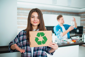 Young family sorts materials in the kitchen for recycling. Recyclable materials must be separated. Wife is holding a cardboard box with a green recycling sign. Husband sorts trash in the background