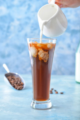 Pouring of milk into cold coffee in glass on color background