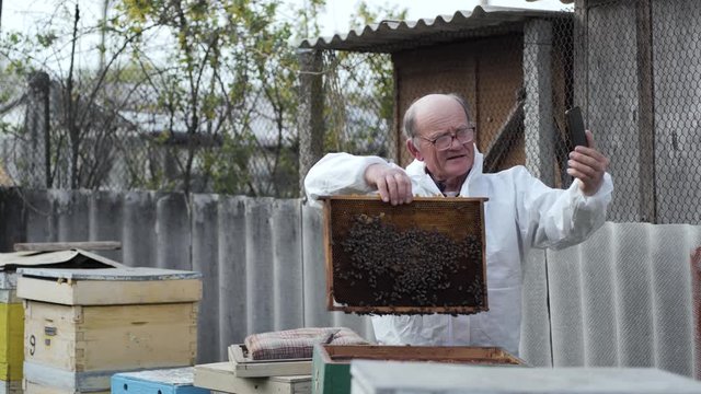 elderly male influencer in protective suit with glasses for sight makes video blog for followers with mobile phone in hands shows honeycomb on background of honey bees standing in an apiary
