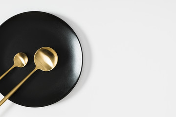 Kitchen concept. Black empty ceramic plate and golden cutlery on white background. Flat lay, top view, copy space