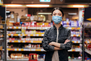 A young beautiful Asian woman is inside a supermarket, shopping for groceries, while wearing a medical mask. Communicate about health and safety measures in commonly used spaces, it times of pandemic