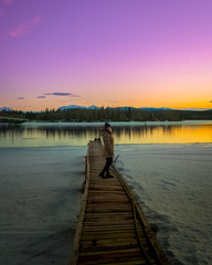 Obraz premium Woman standing on a pier, boat launch with snow & purple, orange and pink stunning sunset with mountains and river, lake reflection. 
