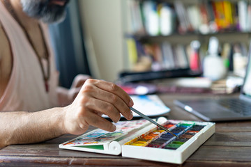 Fototapeta premium man artist painting with paint brush and watercolor palette on paper in front of laptop, closeup. Selective focus