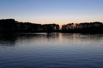 The surface of the water with ripples and reflections of sunset on background of a calm symmetrical landscape in a light misty haze, soft focus.