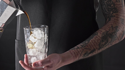 A man with a tattoo pours coffee in a glass with ice around a dark background. 4K UHD video, 2160p.