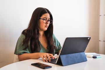 Young spanish brunette wearing glasses teleworking with her hybrid tablet laptop adapting her business to the new normal after the covid 19 global pandemic. She looks concentrated