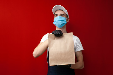 young guy food delivery man in a protective mask and gloves holds a mock up of an empty bag, a man holds a parcel during coronavirus, template