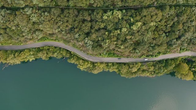 Flying along a road closed to Como Lake, Italy. Aerial view of a forest road with a car moving among green trees and blue water of lake.