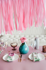 Birthday table setting in pink and colors with rose in vase. Streamers garland background. Baby shower or girl party.