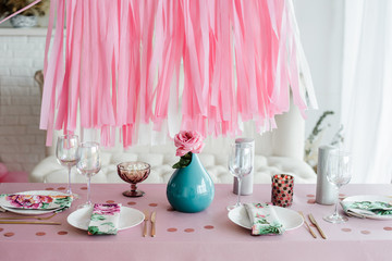 Birthday table setting in pink and colors with rose in vase. Streamers garland background. Baby shower, girl party decoration.
