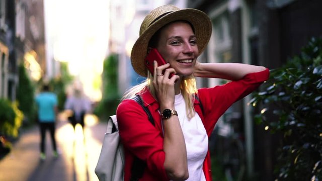 Happy millennial female traveller dressed in casual look smiling during smartphone conversation, cheerful hipster girl in hat walking around city streets with cellular phone calling via application 

