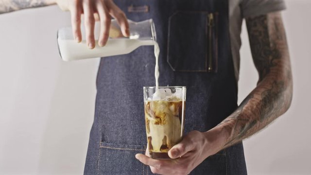 Barista's hand pours fresh milk into glass with ice coffee.