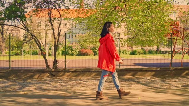 side view brunette in casual shoes and red raincoat walking in residential area slow motion on the background playground spring season in sunlight