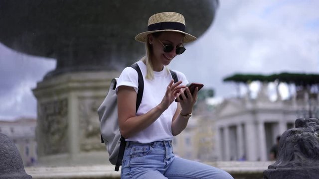 Happy hipster girl in sunglasses enjoying rest near fountain typing text message in chat on smartphone. Female using 4g roaming internet on cellular phone for share photos in social network via app
