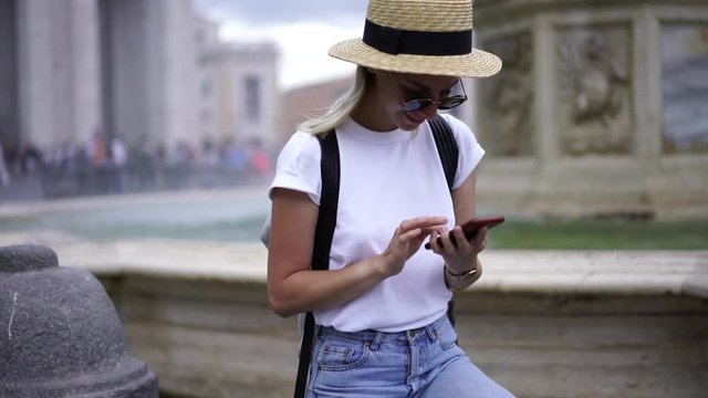 Happy hipster girl enjoying rest near fountain typing text message in chat on smartphone. Stylish female tourist using 4g roaming internet on cellular phone for share photos in social network via app

