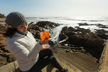 Asian mixed-race woman sitting with a cocktail on the sea rocks on sunny day.