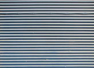 Background of a closed iron curtain painted blue. Horizontal lines of light blue painted metal enclosure illuminated by sunlight.       