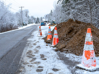 Orange cones warn drivers of a utilities work zone, as a crew and backhoe work on trenches in the ground next to a residential city street, on a frosty winter day in Bemidji, Minnesota.
