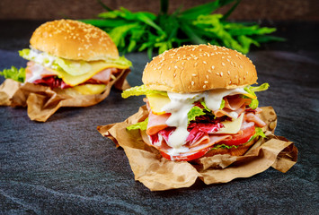 Two sandwiches with ham, cheese, tomatoes, lettuce on dark background.