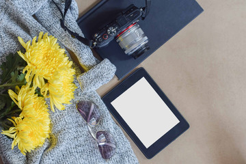 tabletop view of travel concept with a tablet with a blank screen, shades, camera, and black notebook on a plaid cardigan sweater and yellow flowers with copy space.
