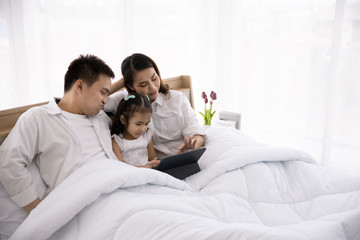 Happy Asian family using technologies for fun. Father, mother and daughter looking at digital tablet on the bed in the home