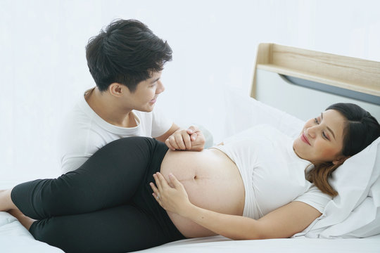 Asian pregnant woman and her husband laying on the bed while spending time together.