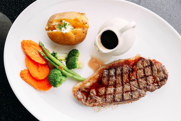 Top view of Charcoal grilled boneless dry-aged Top Loin steak served with BBQ sauce, stir-fried vegetables and baked potato in a white plate.