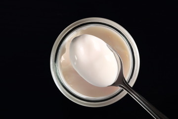 Spoon with yogurt on open small glass jar at black background. D