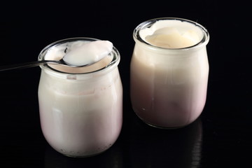 Spoon with yogurt on open small glass jar at black background