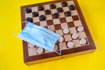 Board game wooden checkers and a medical mask on a yellow background, the concept of leisure during the quarantine