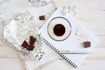 A cup of coffee, chocolate, a sheet of paper and a pen on a white wooden surface. Background for work at home in quarantine. The difficulty of adapting work at home.