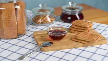 A vase of cherry jam and a stack of shortbread cookies on a wooden tray stand on a table with a white tablecloth in a cage