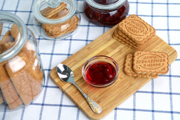 A vase of cherry jam and a stack of shortbread cookies on a wooden tray stand on a table with a white tablecloth in a cage. View from above