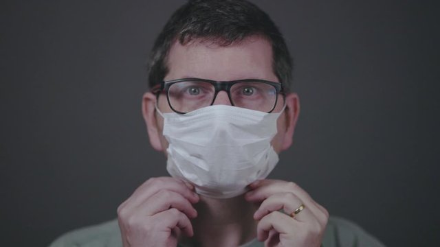 Caucasian male with blue eyes and glasses on looking straight into the camera putting a surgical medical face mask on, securing it correctly covering mouth and nose. 60fps clip in normal speed.