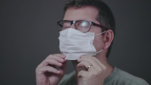 Sideways view of caucasian male with glasses putting on a surgical medical face mask, securing it correctly covering mouth and nose and taking it off. Studio shot 60fps clip in normal speed.