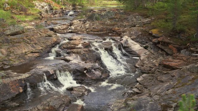The waters of the shallow river gently cascade over the rocky riverbed. Moss and lichen cover the river banks.