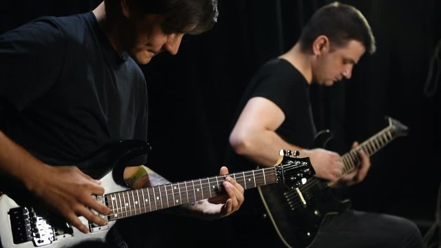 Two male guitarists in black clothes performing song in dark room.