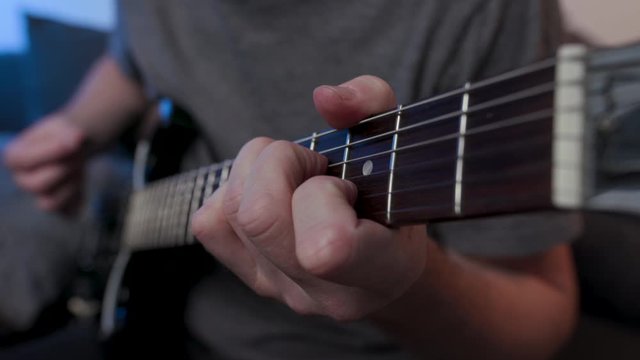 Man practices chords on electric guitar at home sitting on couch, side on view from guitar neck.