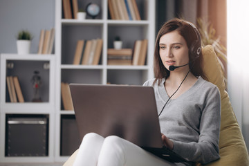 Concentrated young girl working on laptop in headset while sitting on yellow bag chair. Beautiful woman with dark hair using computer for remote work at home